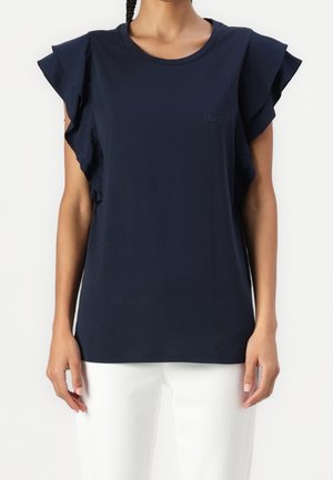 Kobieta ubrana w granatowy top z falbankami na rękawach i białe spodnie, stojąca na jednolitym białym tle.