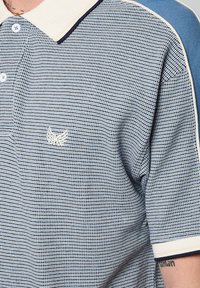 Polo à manches courtes de couleur bleu clair avec un motif à carreaux, col blanc contrastant et petit logo sur la poitrine. Tissu texturé.