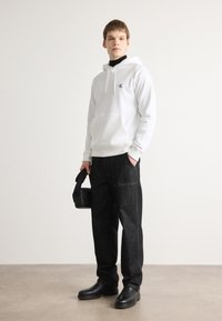 Sudadera blanca con cordones y un logo, combinada con pantalones de mezclilla oscuros. Calzado negro y un pequeño bolso negro completan el look.