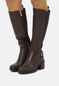 Botas de cuero marrón hasta la rodilla con cremallera lateral y acentos en hardware dorado, que presentan un tacón grueso y suela de goma negra texturizada.