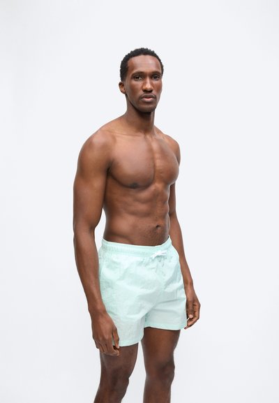 Φορώντας ανοιχτό μπλε μαγιό, ένας γυμνασμένος άνδρας στέκεται χωρίς μπλούζα με ουδέτερη έκφραση, μπροστά σε λευκό φόντο.