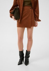 Mini gonna in corduroy marrone con chiusura laterale a bottoni, indossata con una giacca in corduroy marrone oversized e stivaletti neri con punta.