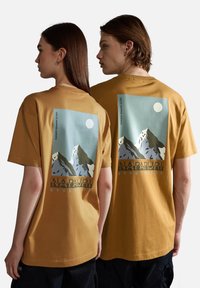 Dos personas vestidas con camisetas mostaza que tienen un gráfico de montaña, una luna y el texto "NAPAPIJRI geographic" en la parte posterior, de pie una al lado de la otra.