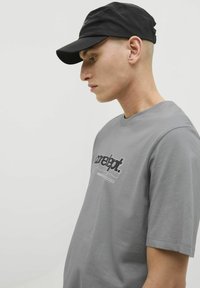Grå T-shirt med rund hals og korte ærmer, med et sort-hvidt grafisk print. Båret med en sort kasket. Glat bomuldsstruktur.