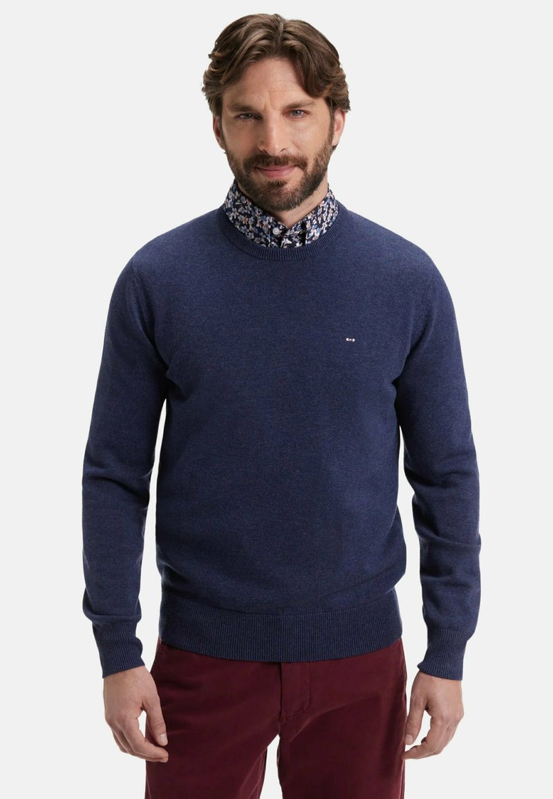Pull en maille bleu marine avec des poignets et un ourlet côtelés, col rond, porté par-dessus une chemise à col à fleurs ; présente un petit logo sur la poitrine.