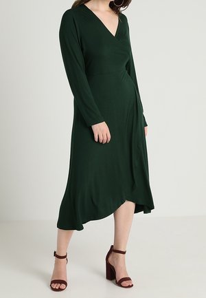 Kobieta ubrana w ciemnozieloną sukienkę kopertową z długim rękawem i asymetrycznym dołem, do której dobrano bordowe sandały na słupku z paskiem wokół kostki.