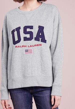 Person, die einen hellgrauen Sweatshirt mit großem dunkelblauem "USA", rotem "Ralph Lauren" und einem kleinen amerikanischen Flaggen-Patch vorne trägt.