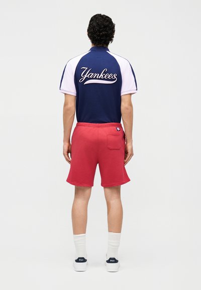 Άτομο με σγουρά μαλλιά που φοράει μπλε σκούρο και λευκό t-shirt των Yankees, κόκκινα σορτς, λευκές κάλτσες και λευκά παπούτσια Polo, στέκεται γυρισμένο με την πλάτη.