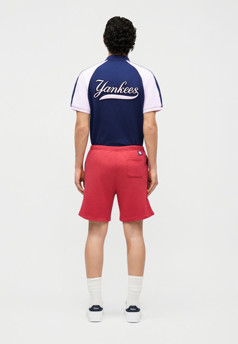 Persoon met krullend haar die een marineblauw-witte Yankees-shirt, rode korte broek, witte sokken en witte Polo-sneakers draagt, staand met de rug naar de kijker.