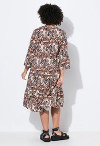 Beige, bruine en zwarte jurk met bloemenpatroon tot op de knie, met driekwart mouwen en een gathering in de taille, gedragen met zwarte sandalen.