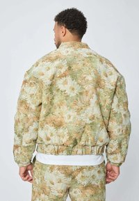 Homme de dos portant un ensemble assorti en tissu à motifs floraux, composé d'une veste et d'un pantalon en tons beige, vert et blanc, sur une chemise blanche.