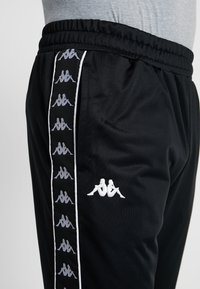 Svarta sportiga joggingbyxor med elastisk midja, med en vit sidostrand med upprepade logotyper och en liten logotyp på låret.