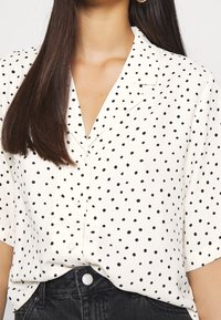 Blusa de manga corta en tela blanca con lunares negros, con cuello con muesca y cierre de botones, combinada con denim oscuro.