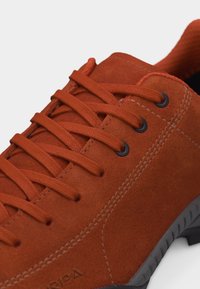 Αθλητικό παπούτσι από σουέντ σε σκουριά χρώμα με κορδόνια στο ίδιο χρώμα, μαύρα μεταλλικά κρόσσια, ορατές ραφές και γκρι εξωτερική στρώση στην σόλα από καουτσούκ.