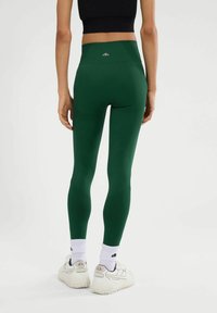 Leggings verdes con cintura alta, fabricados con material elástico, que presentan acentos texturizados y un logo en la parte posterior. Llevados con zapatillas blancas.