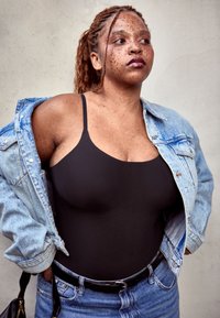 Mlada ženska z pegicami in spetimi lasmi nosi črno majico, jeans jakno in hlače ter stoji s rokami na bokih ob enobarvni steni.