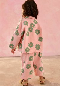 Rosa bomullsoutfit bestående av en lös jacka och matchande byxor med stora gröna blommönster och orange centra. Designad för barn.