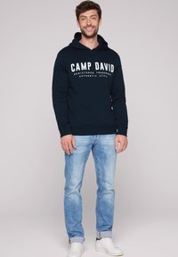Granatowy sweatshirt z białym napisem "CAMP DAVID", długie rękawy i kieszeń kangurzą, w zestawieniu z jasnoniebieskimi dżinsami i białymi trampkami.