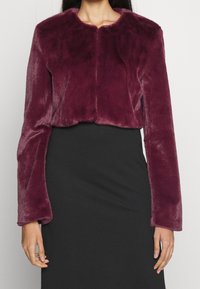 Veste courte en fausse fourrure bordeaux avec manches longues, col rond et texture douce, portée sur une robe noire ajustée.