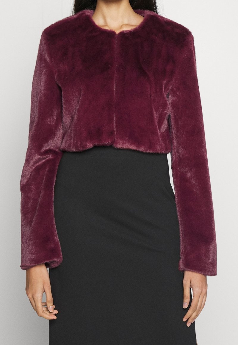 Veste courte en fausse fourrure bordeaux avec manches longues, col rond et texture douce, portée sur une robe noire ajustée.