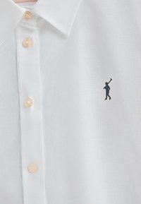 Una camicia bianca con bottoni, realizzata in tessuto testurizzato, con bottoni chiari e una piccola figura ricamata in filo scuro sul lato sinistro del petto.
