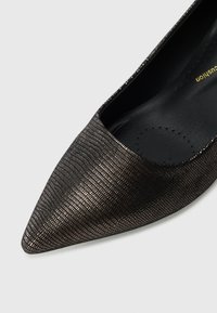 Pumpe med spiss tå i svart med en teksturert metallisk finish. Har et glatt interiør og et lavt snitt for et elegant utseende.