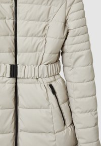 Giacca imbottita beige con design trapuntato, chiusura a zip, tasche laterali e vita con cintura. Presenta una texture liscia e accessori neri.