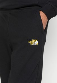 Černé tepláky s hladkým povrchem, s kapsou a žlutým logem "The North Face" na stehně.