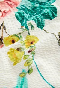 Teksturirana bela tkanina z natisnjenim cvetličnim vzorcem, ki vključuje rumene cvetove, zelene liste ter pridih rožnate in rjave barve na cvetnih listih.
