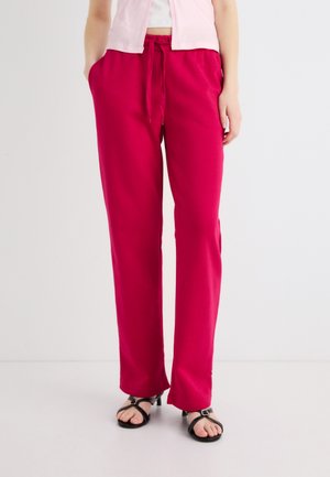 Donna che indossa pantaloni rosa acceso con coulisse, sandali neri aperti con tacco e top corto rosa chiaro su sfondo bianco.