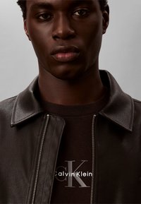 Νέος άνδρας που φοράει σκούρο καφέ δερμάτινο μπουφάν πάνω από μαύρο πουκάμισο Calvin Klein με μεγάλο λογότυπο "CK" μπροστά.