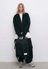 Svart tygryggsäck med meshpaneler och dragkedjestängning, med topphandtag och avtagbara remmar. Bärs av en person i en svart outfit.