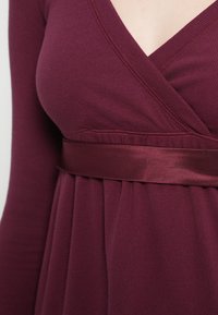 Μπορντό φόρεμα με βαθύ V-λαιμόκοψη, ριμπ υφή και σατέν ζωνάκι στη μέση. Διαθέτε�ι πιέτες στη μέση, που δημιουργούν μια εφαρμοστή σιλουέτα.