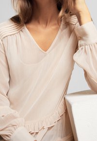 Femme portant un chemisier léger beige clair, transparent, à manches longues, avec des détails plissés et un galon à volants, reposant son bras sur une surface blanche.