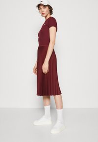Abito plissettato bordeaux con maniche corte, tessuto strutturato; abbinato a sneakers bianche e calze, completato da un cappellino bianco.