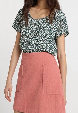 Vrouw draagt een groen bloemenprint kort mouwen top, ingestopt in een hoog uitgesneden roze corduroy rok met twee steekzakken aan de voorkant.