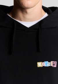 Juodas džemperis su gobtuvu ir priekinė kišene, puoštas daugiabriauniu "LEGACY" siuvinėjimu. Medžiaga atrodo minkšta ir tekstūruota. Raišteliai yra juodos spalvos.