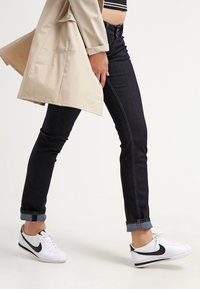 Jeans ajustados negros con un ligero brillo, doblados en los tobillos, combinados con zapatillas Nike blancas con un swoosh negro. Abrigo trench beige.