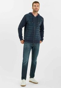 Chaqueta acolchada azul marino con capucha y acentos a rayas rojas y blancas, combinada con jeans azul oscuro y zapatillas blancas con suelas color beige.