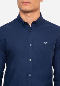 Camisa de botones azul marino en tejido de algodón con cuello puntiagudo, que presenta un pequeño emblema bordado en el lado izquierdo del pecho y puños estándar.