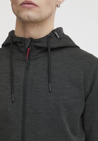 Tmavohnedá mikina na zips s textúrovým povrchom, s kapucňou na šnúrku, červeným zipsom a brandingom na krku.