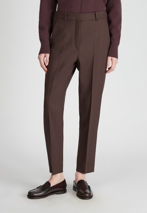 Persoon in een aansluitende bruine broek, donkere leren loafers, en een paarse trui met lange mouwen, staand tegen een effen lichte achtergrond.