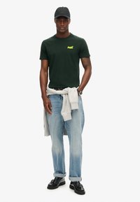 Man wearing dark green t-shirt, light blue jeans with cuffs, black shoes, black cap, and gray sweatshirt tied around waist.