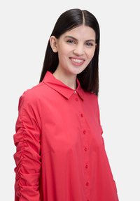 Chemise rouge à boutons avec un col, présentant des détails froncés le long des manches et un tissu lisse et léger.