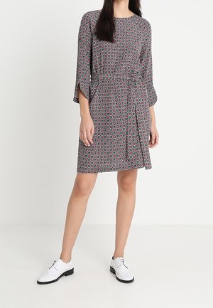 Woman wearing a knee-length patterned dress with three-quarter sleeves and white lace-up shoes, standing against a plain white background.