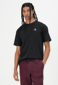 Schwarzes Kurzarm-T-Shirt mit einem dreieckigen Logo auf der linken Brust, getragen mit bordeauxfarbenen Cordhosen. Glatte Textur, lockerer Schnitt.