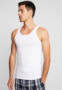 White ribbed tank top featuring wide shoulder straps, paired with plaid pajama shorts. Fabric appears soft and breathable.