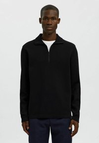 Zwarte pullover met een half-rits kraag, lange mouwen en een gestructureerde stof. Gekozen over een wit T-shirt, gecombineerd met donkere broeken.