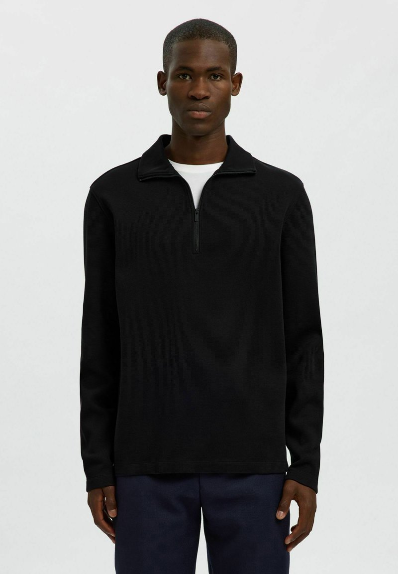 Zwarte pullover met een half-rits kraag, lange mouwen en een gestructureerde stof. Gekozen over een wit T-shirt, gecombineerd met donkere broeken.