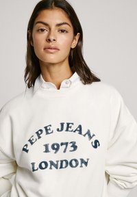 Sudadera blanca con un estampado redondo en azul marino que dice "PEPE JEANS 1973 LONDRES," llevada sobre una camisa blanca de cuello, presenta un ajuste relajado.
